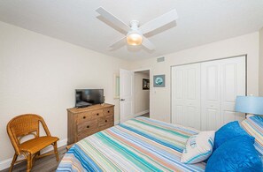 Une chambre à coucher avec un lit, un téléviseur posé sur une commode, une chaise, une porte et un ventilateur de plafond.