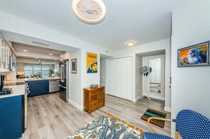 Une cuisine moderne avec un îlot central, un coin repas agrémenté d’un tapis à motifs, et un couloir avec une porte et un ventilateur.