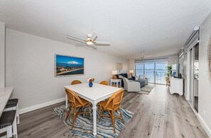 A modern living room with a dining area, a ceiling fan, a large window, and a view of the sea.
