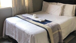 Un lit bien fait, avec du linge de lit blanc et un élément décoratif bleu, une fenêtre avec des rideaux et un sol à carreaux.