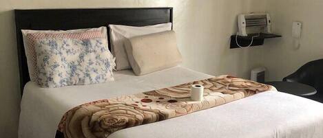 Una cama bien tendida con un cojín floral, una manta color beige y una colcha blanca. Se observa un cabecero negro y una unidad de aire acondicionado fija en la pared a la derecha.