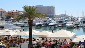 Un puerto deportivo con barcos amarrados, un hotel al fondo y zonas de estar al aire libre con sombrillas.