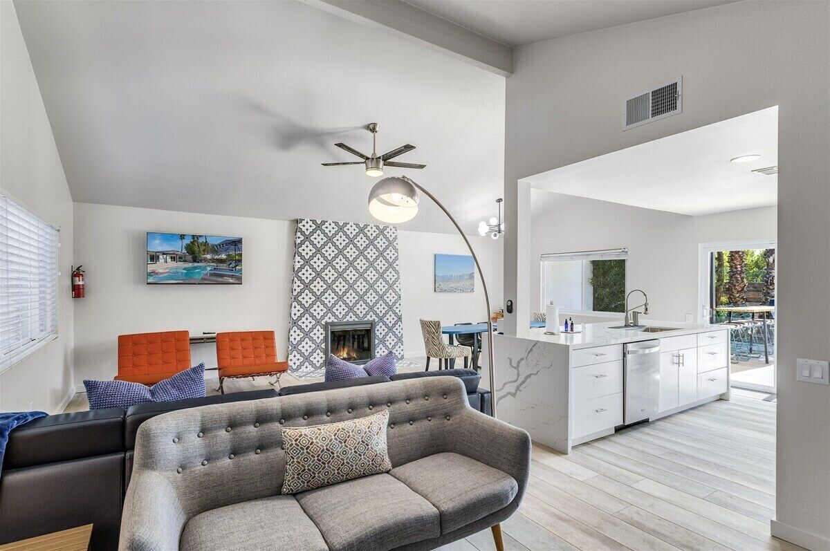 A modern living room with a grey sofa, orange chairs, a fireplace, and a kitchen area with white cabinets and a large window.