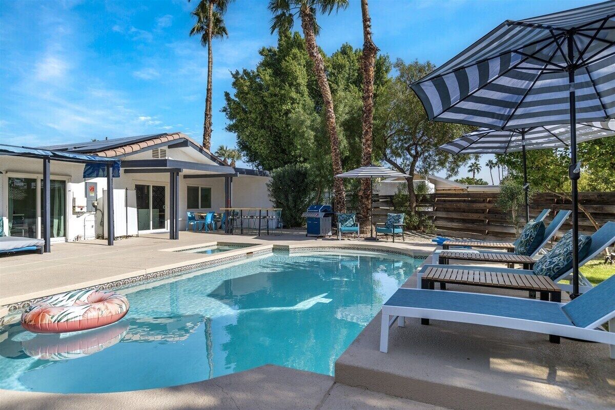 Une piscine dotée de chaises longues et de parasols, entourée de palmiers et d’un patio couvert.