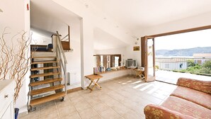 A spacious living area with a staircase, a sofa, a television, and a balcony with a view of the hills.