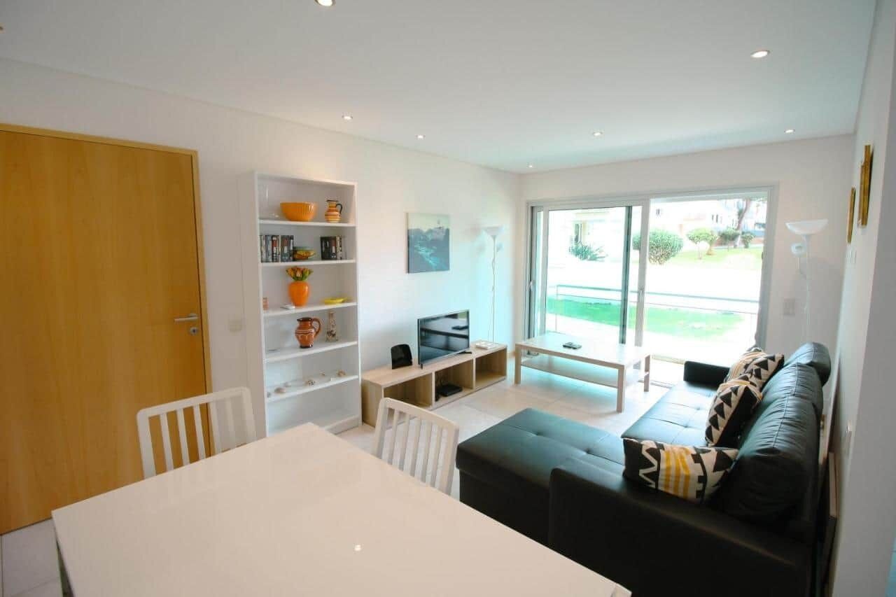 Een moderne woonkamer met een eethoek, een boekenkast, een televisie en een grote schuifpui die uitkomt op een tuin.