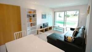 Een moderne woonkamer met een eethoek, een boekenkast, een televisie en een grote schuifpui die uitkomt op een tuin.