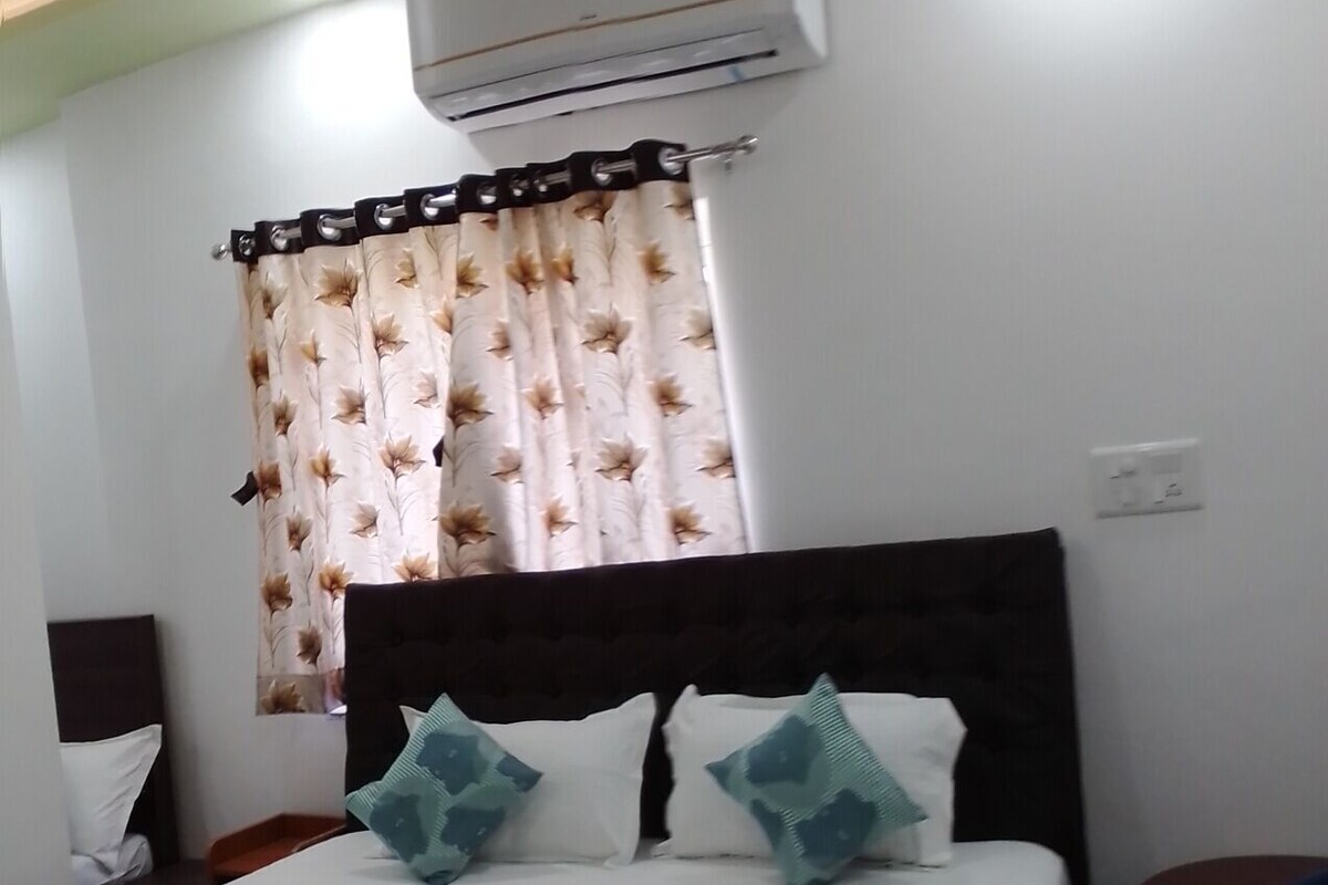 A hotel room with a bed, a window with curtains, an air conditioner, and a small table.