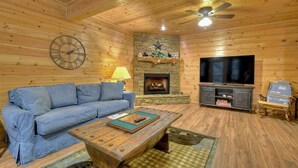 A cozy living room with a blue sofa, a wooden coffee table, a stone fireplace, and a television on a wooden stand. - Stoney Creek Retreat - Cherry Log, GA (Cherry Log)
