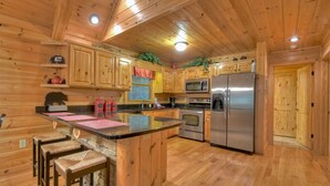 A wooden cabin interior with a kitchen featuring wooden cabinets, a stainless steel refrigerator, and a bar area with stools. - Stoney Creek Retreat - Cherry Log, GA (Cherry Log)
