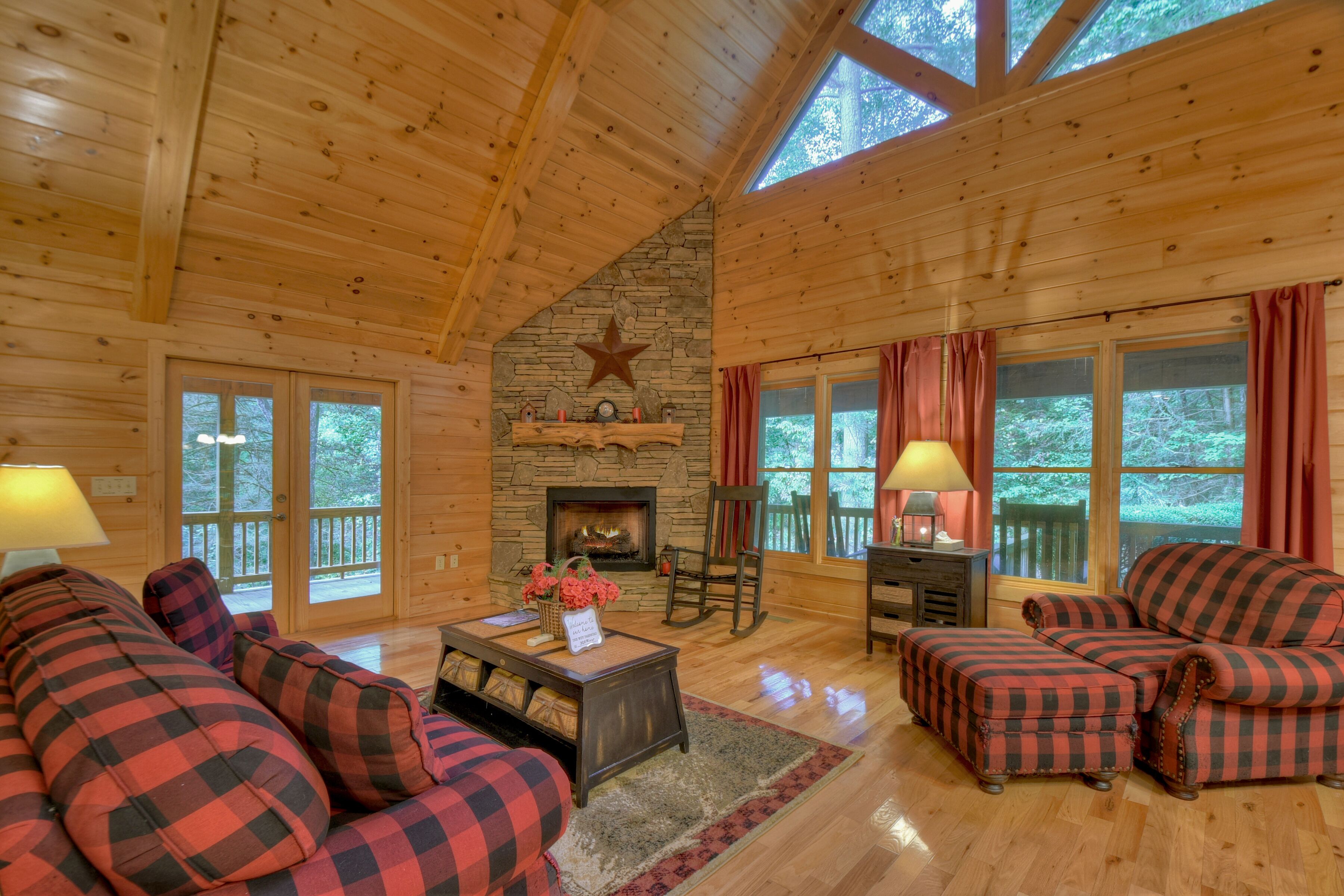 A cozy cabin interior with a stone fireplace, wooden ceiling, and large windows.