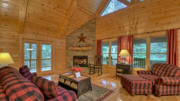 A cozy cabin interior with a stone fireplace, wooden ceiling, and large windows.