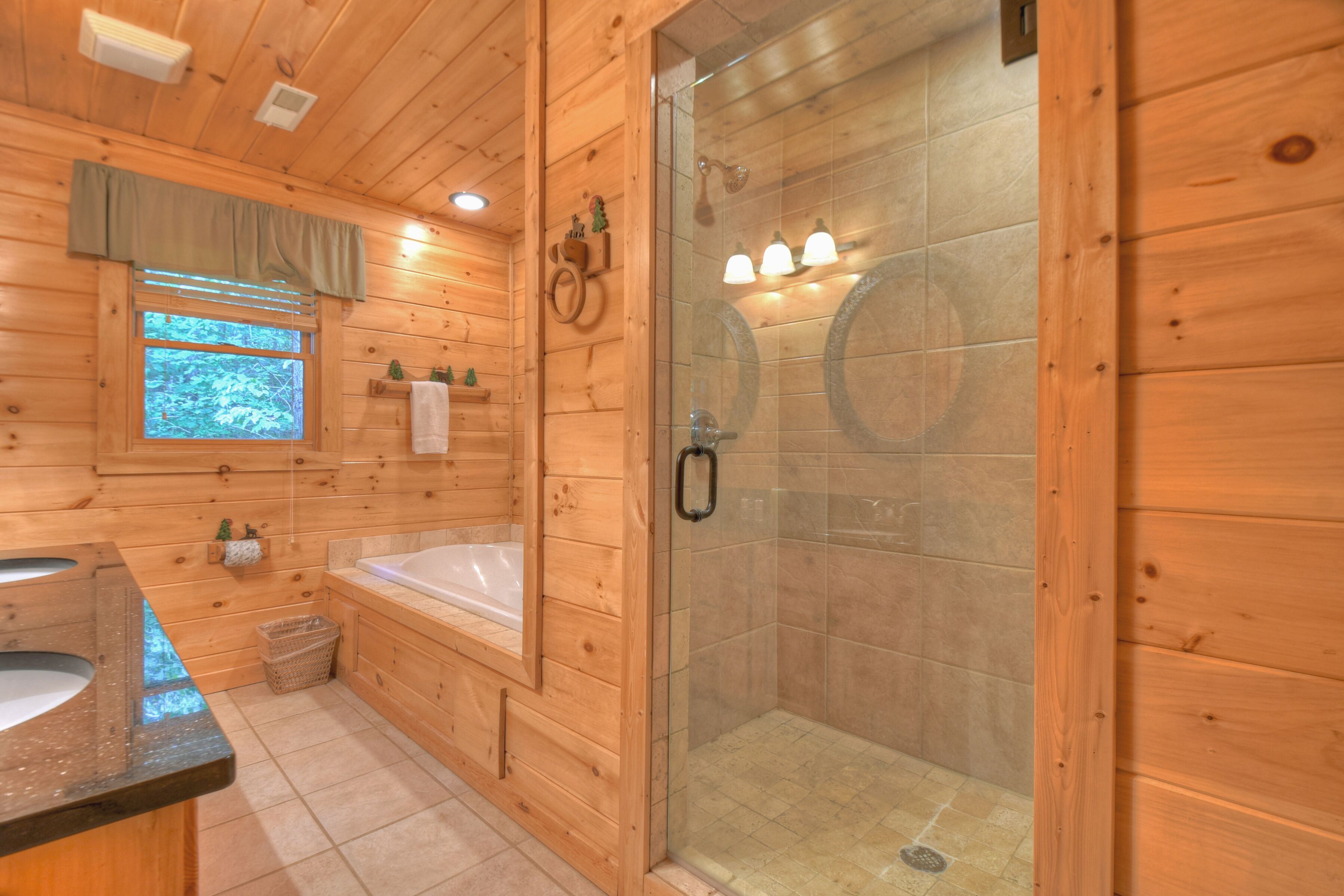 A wooden cabin bathroom with a bathtub, a glass-enclosed shower, and a window with a view of trees.
