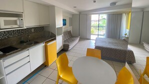 Quarto de hotel com cozinha compacta, uma cama, varanda com vista e área de refeições com cadeiras amarelas.