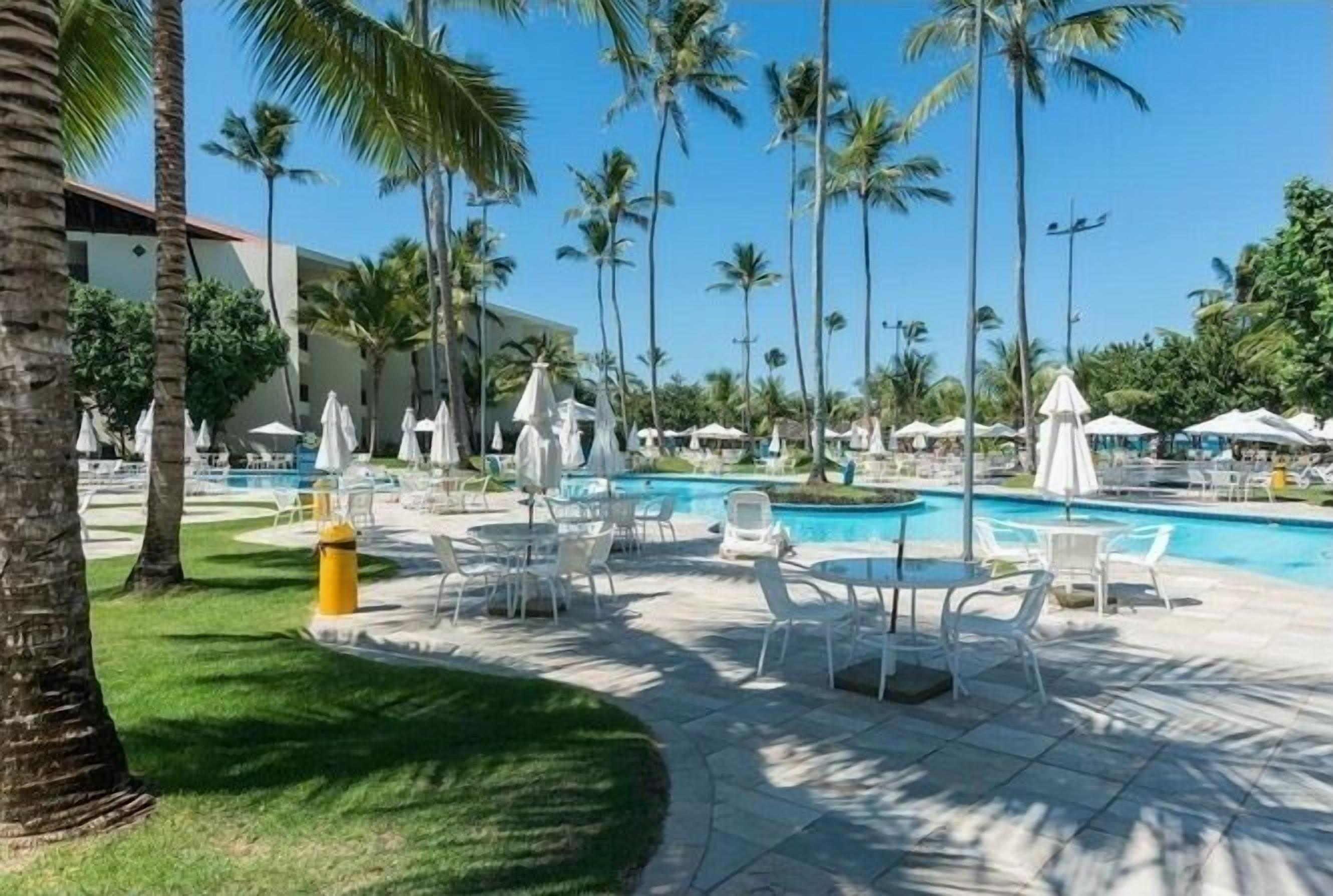 Zona junto a la piscina con sillones blancos, sombrillas y palmeras.