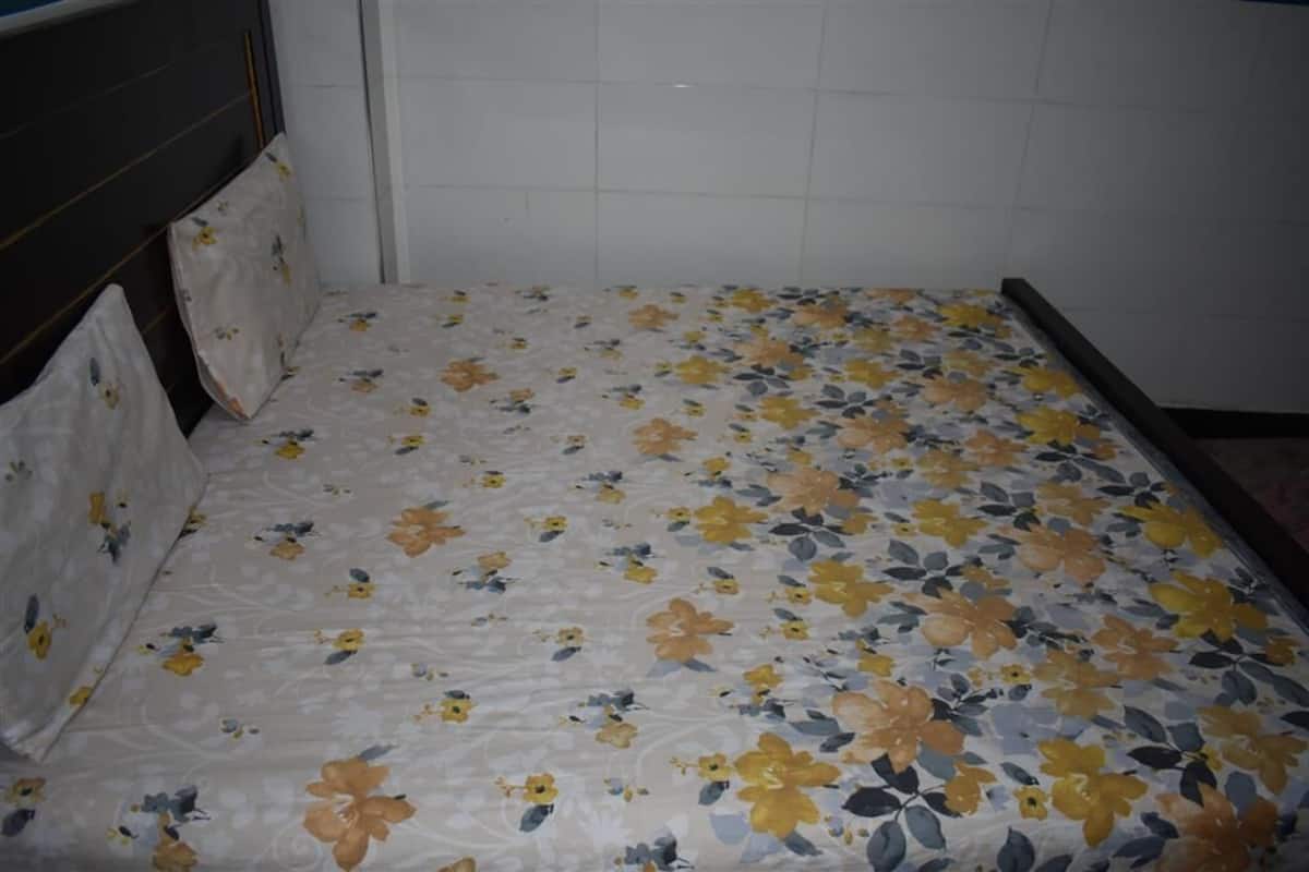 Una habitación con una cama cubierta con ropa de cama estampada con motivos florales, paredes de azulejos blancos y un cabecero oscuro.