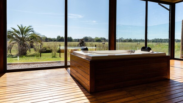 A modern spa room with a large wooden bathtub and a view of a grassy field through large windows. - Terranova Glamping (Luján)