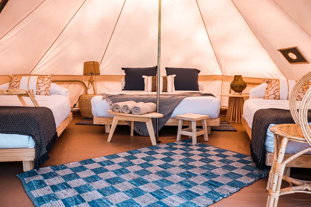 A tented accommodation with two beds, a central table with towels, and a blue checkered rug.