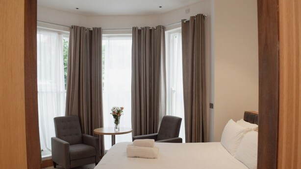 A hotel room with a bed, two armchairs, a small round table with a vase of flowers, and a view of the outdoors through large windows with curtains. - Osborne Apartments (Eastbourne)