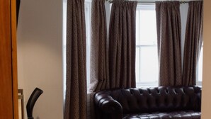 A living room with a dark brown leather sofa, patterned curtains, and a tiled floor. - Osborne Apartments (Eastbourne)