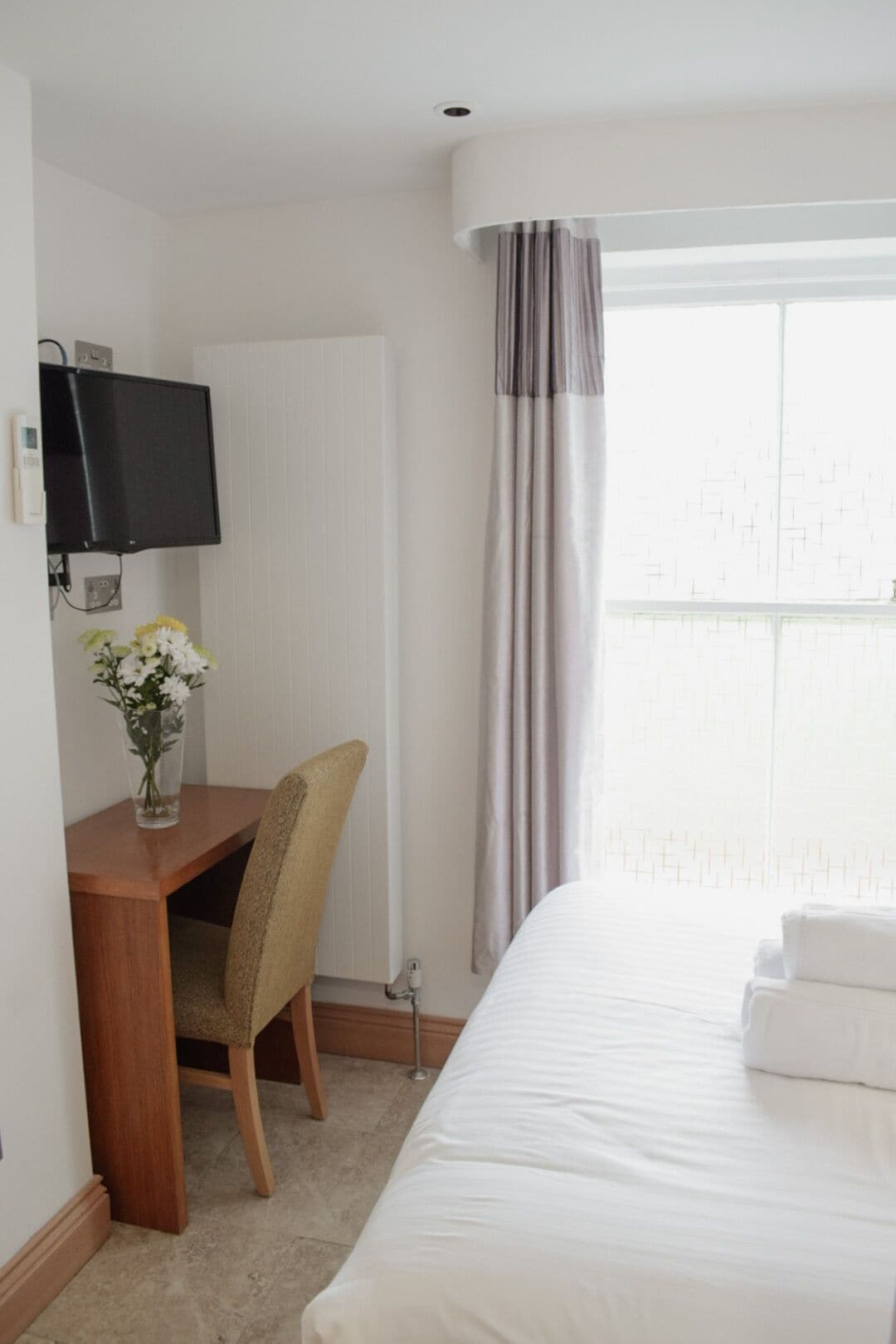 Une chambre d’hôtel comprenant un lit, un bureau avec une chaise, un téléviseur fixé au mur et une fenêtre avec des rideaux.