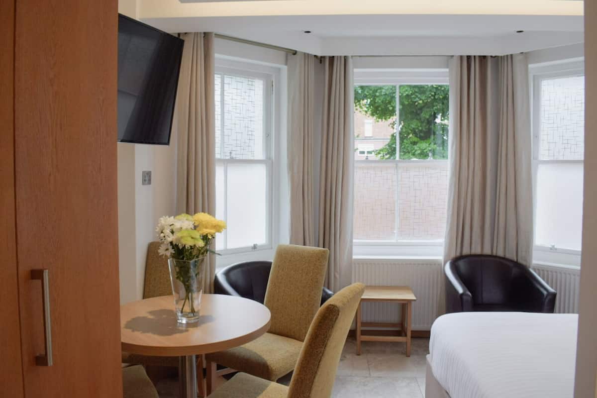 A hotel room with a bed, two chairs, a small table, a TV mounted on the wall, and a window with curtains.