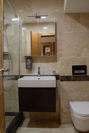 A bathroom with a glass shower enclosure, a white sink with a dark cabinet, a mirror, and a toilet. - Osborne Apartments (Eastbourne)