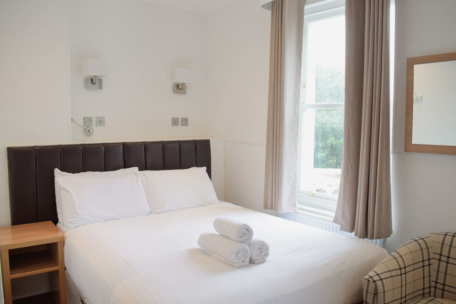 A neatly made bed with white linens and rolled towels, a wooden nightstand, and a plaid armchair.