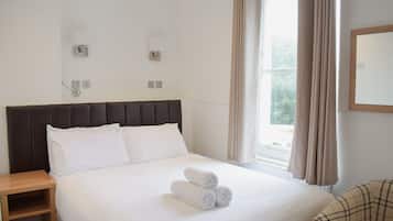 Una cama bien hecha con sábanas blancas y toallas enrolladas, una mesita de noche de madera y un sillón de cuadros.
