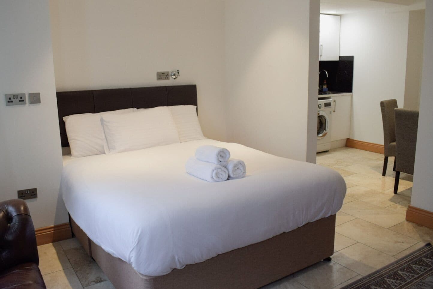 A neatly made bed with white linens and rolled towels, a dark headboard, and a small brown ottoman at the foot of the bed.