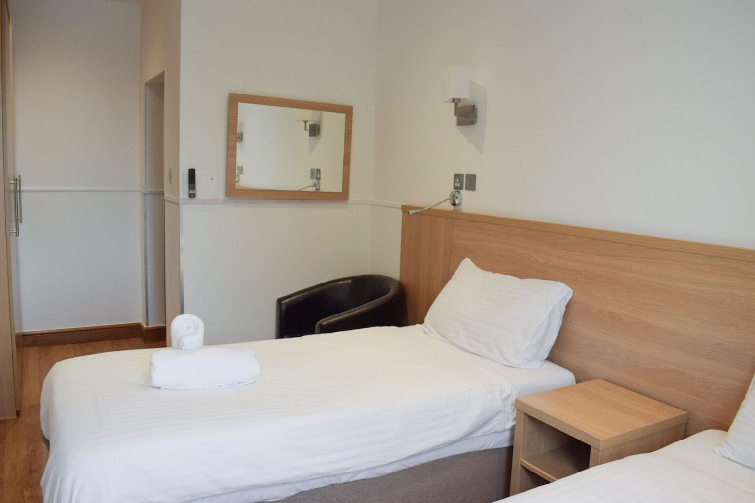 Une chambre d’hôtel avec une tête de lit en bois, un lit simple avec une literie blanche, une table de chevet, une petite table de nuit en bois, une poubelle noire et un miroir fixé au mur.