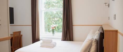 Una cama bien hecha con sábanas y almohadas blancas, una mesita de noche de madera, una silla y una ventana con cortinas.