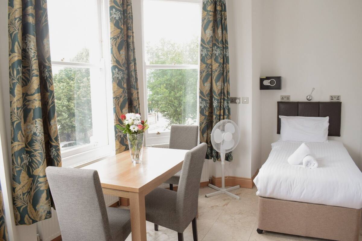 A hotel room with a bed, a bedside table with a vase of flowers, a chair, a standing fan, and a window with curtains.