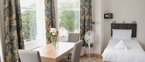 Une chambre d’hôtel avec un lit, une table de chevet ornée d’un vase de fleurs, une chaise, un ventilateur sur pied et une fenêtre avec des rideaux.