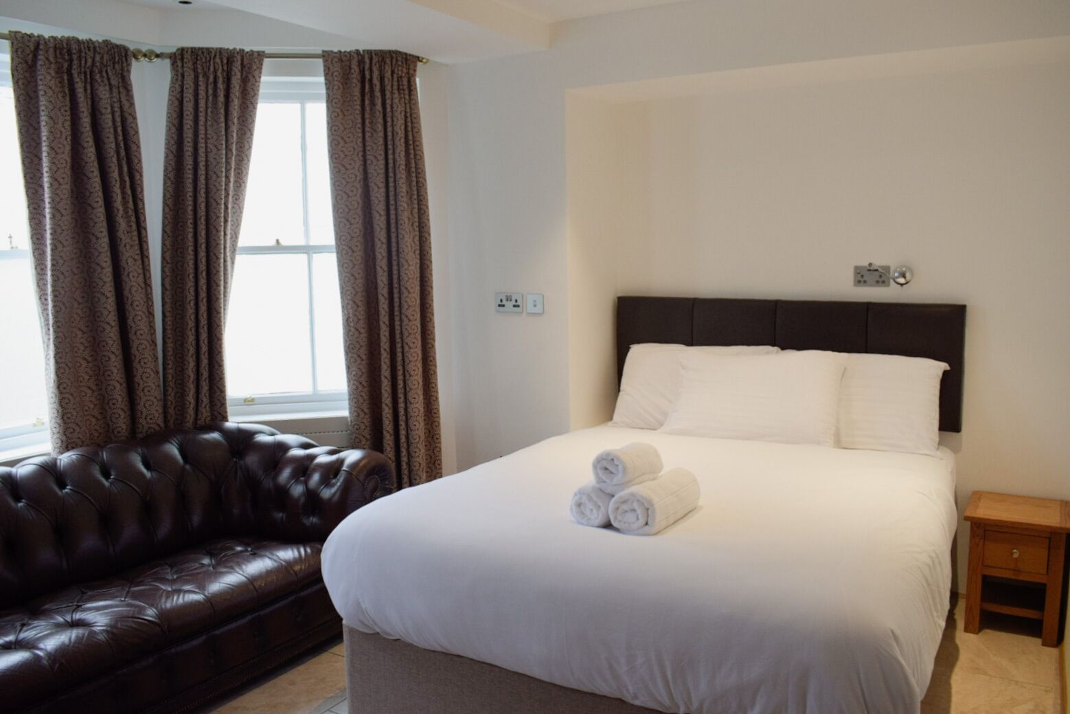 A hotel room with a large bed, a dark brown leather sofa, and a window with curtains.
