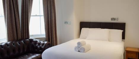 A hotel room with a large bed, a dark brown leather sofa, and a window with curtains.