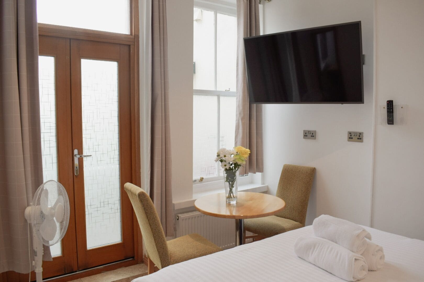 A hotel room with a bed, a chair, a small table with a vase of flowers, a TV on the wall, and a window with curtains.