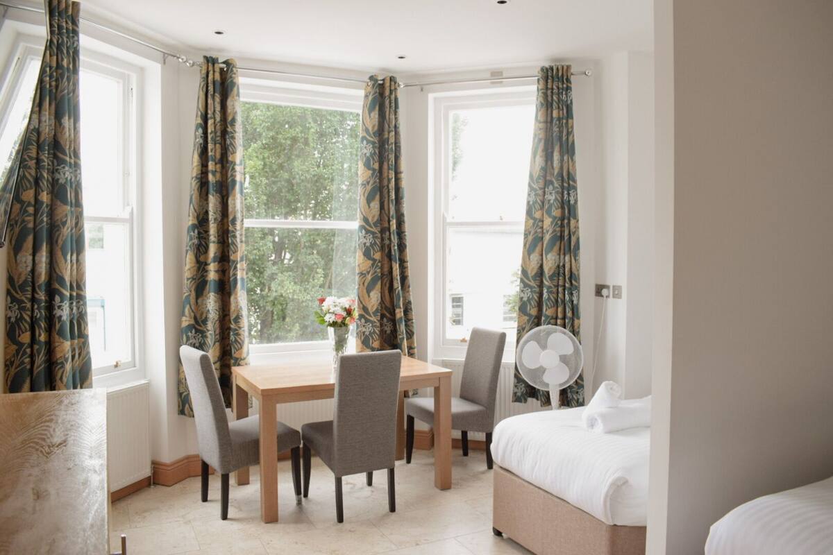 A bedroom with a bed, a table with chairs, a fan, and curtains.