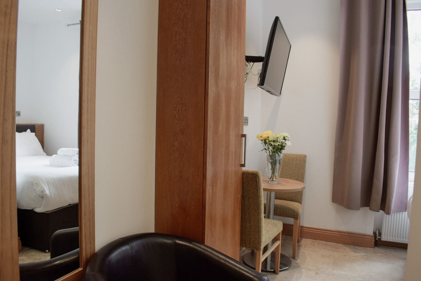 A hotel room with a bed, a TV mounted on the wall, a small table with chairs, and a window with curtains.