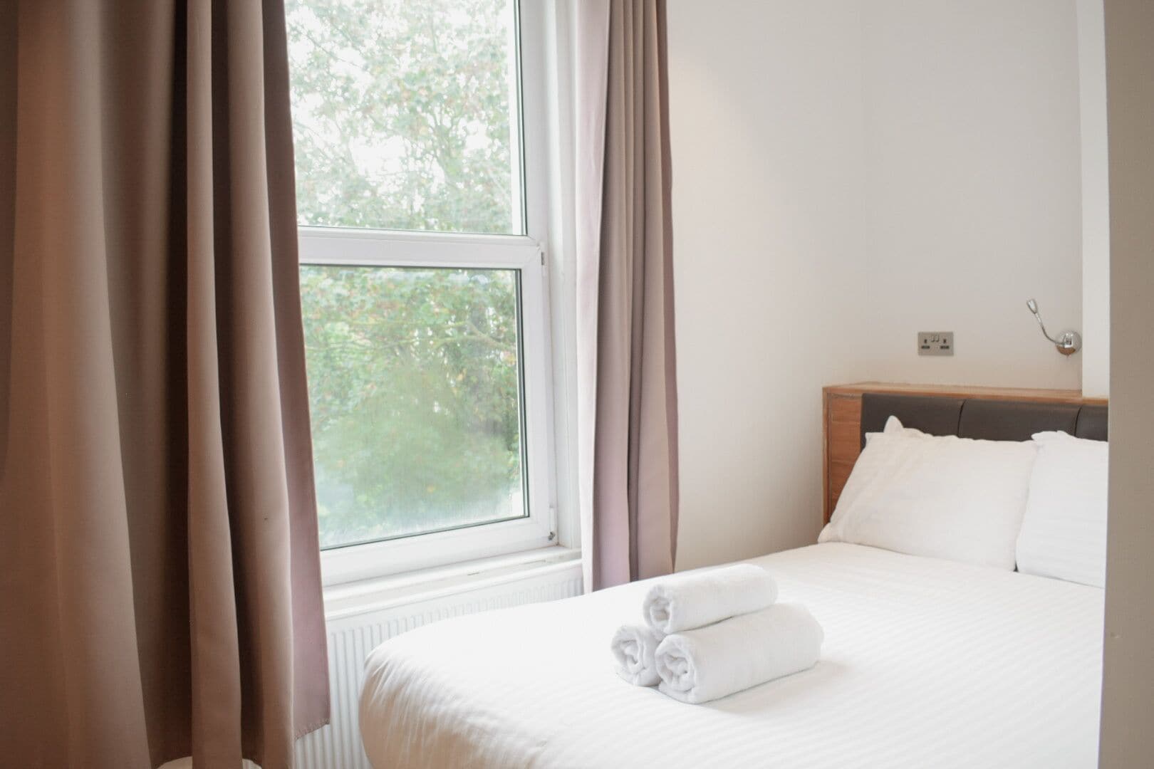 Una habitación de hotel con cama, ventana con cortinas y vista a árboles.