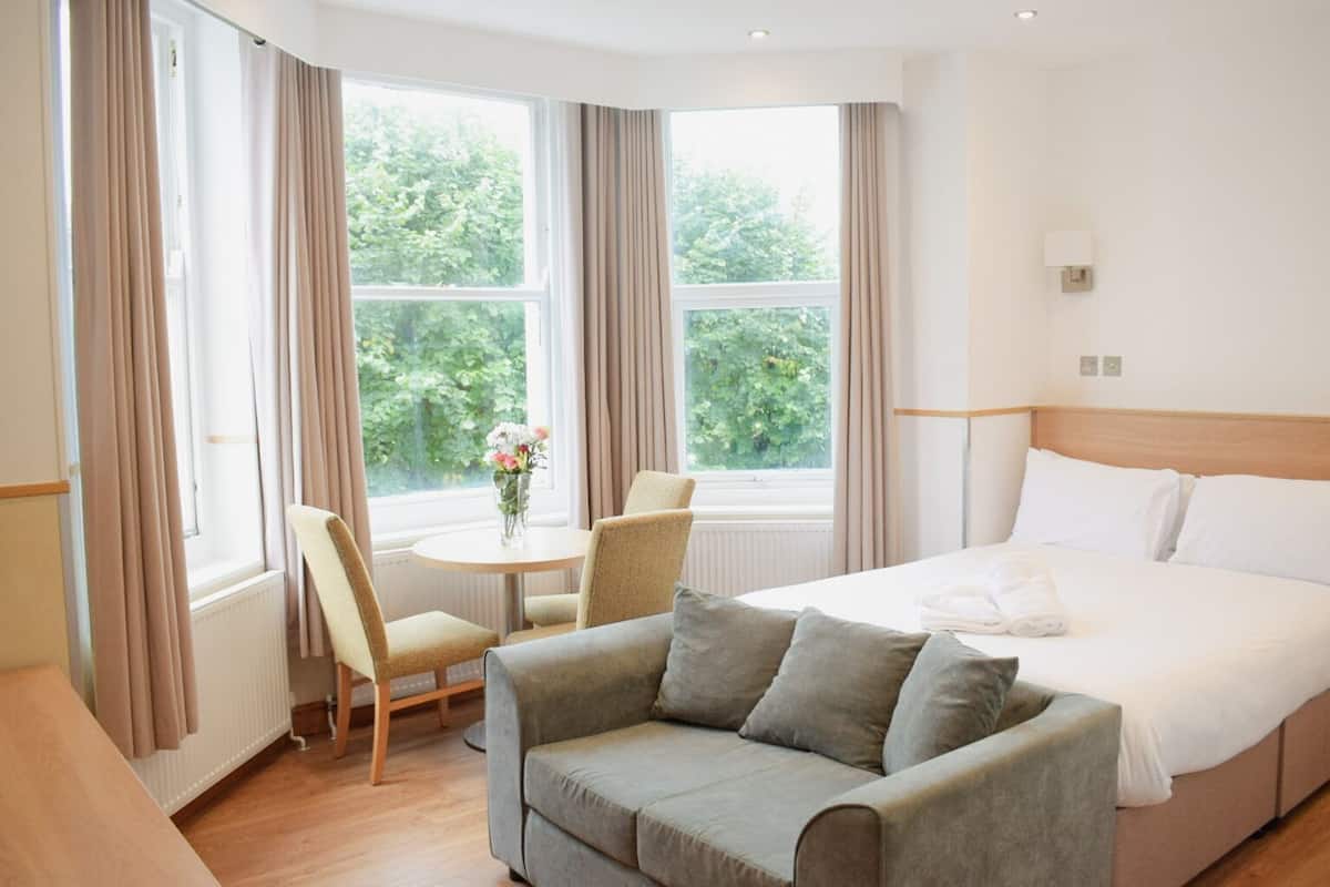 A hotel room with a bed, a sofa, a table with a vase of flowers, and large windows with curtains.
