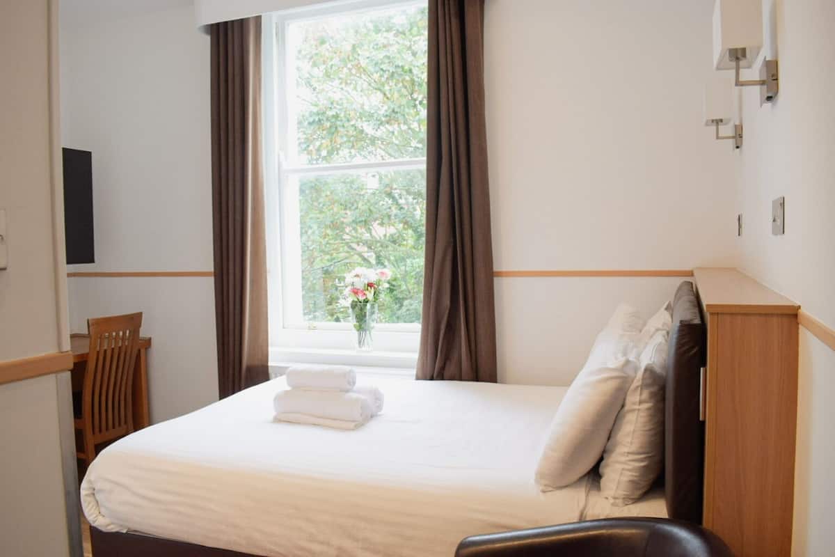A hotel room with a bed, a window with curtains, a chair, and a vase with flowers.