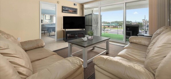 A living room with beige sofas, a flat-screen TV, a small table with a plant, and a sliding glass door leading to a balcony with a view of a parking lot and buildings.