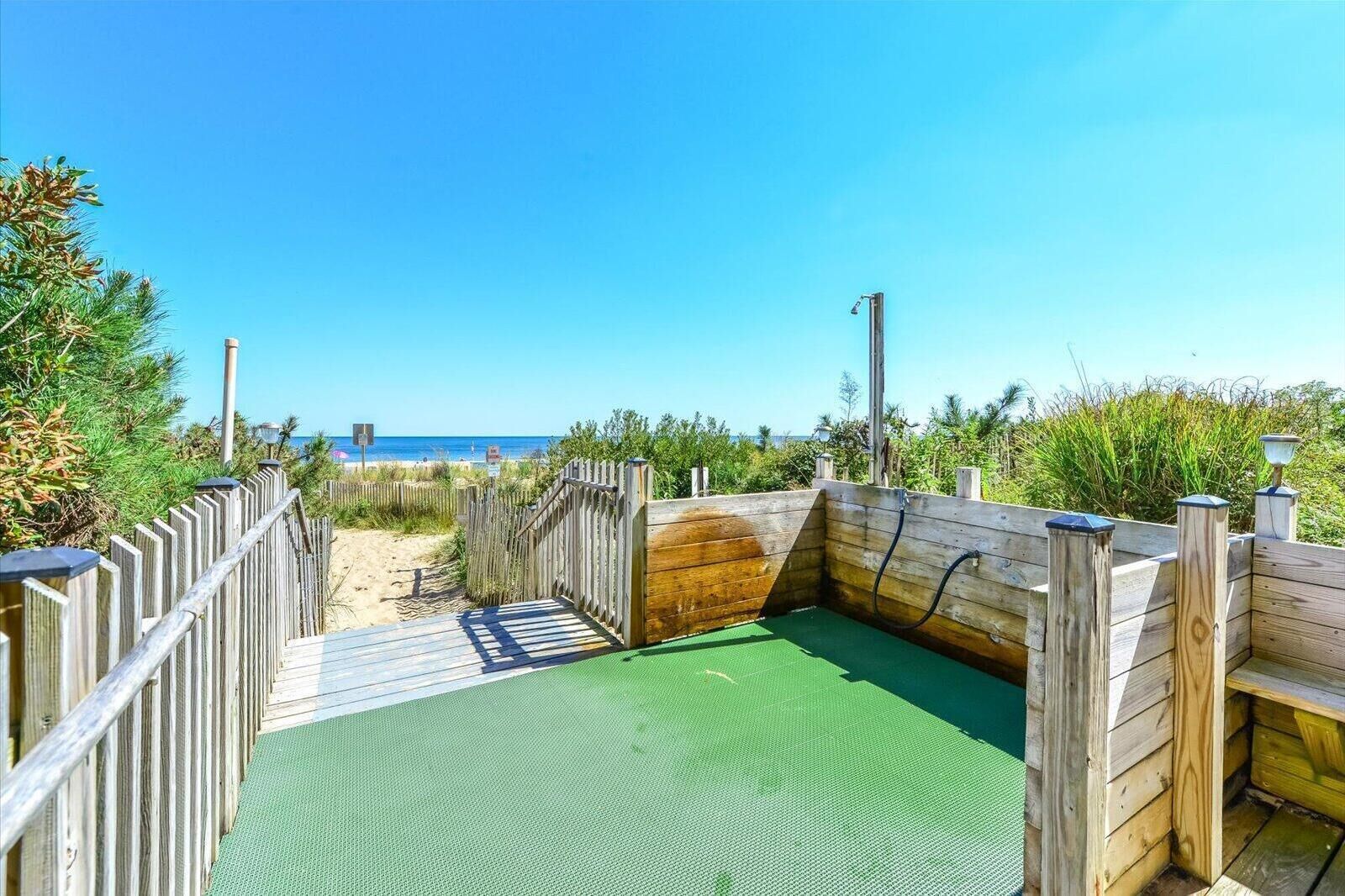 Eine Holzterrasse mit grüner Oberfläche führt zu einem Strandbereich, umgeben von Holzgeländern und Vegetation.