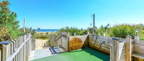 Eine Holzterrasse mit grüner Oberfläche führt zu einem Strandbereich, umgeben von Holzgeländern und Vegetation.