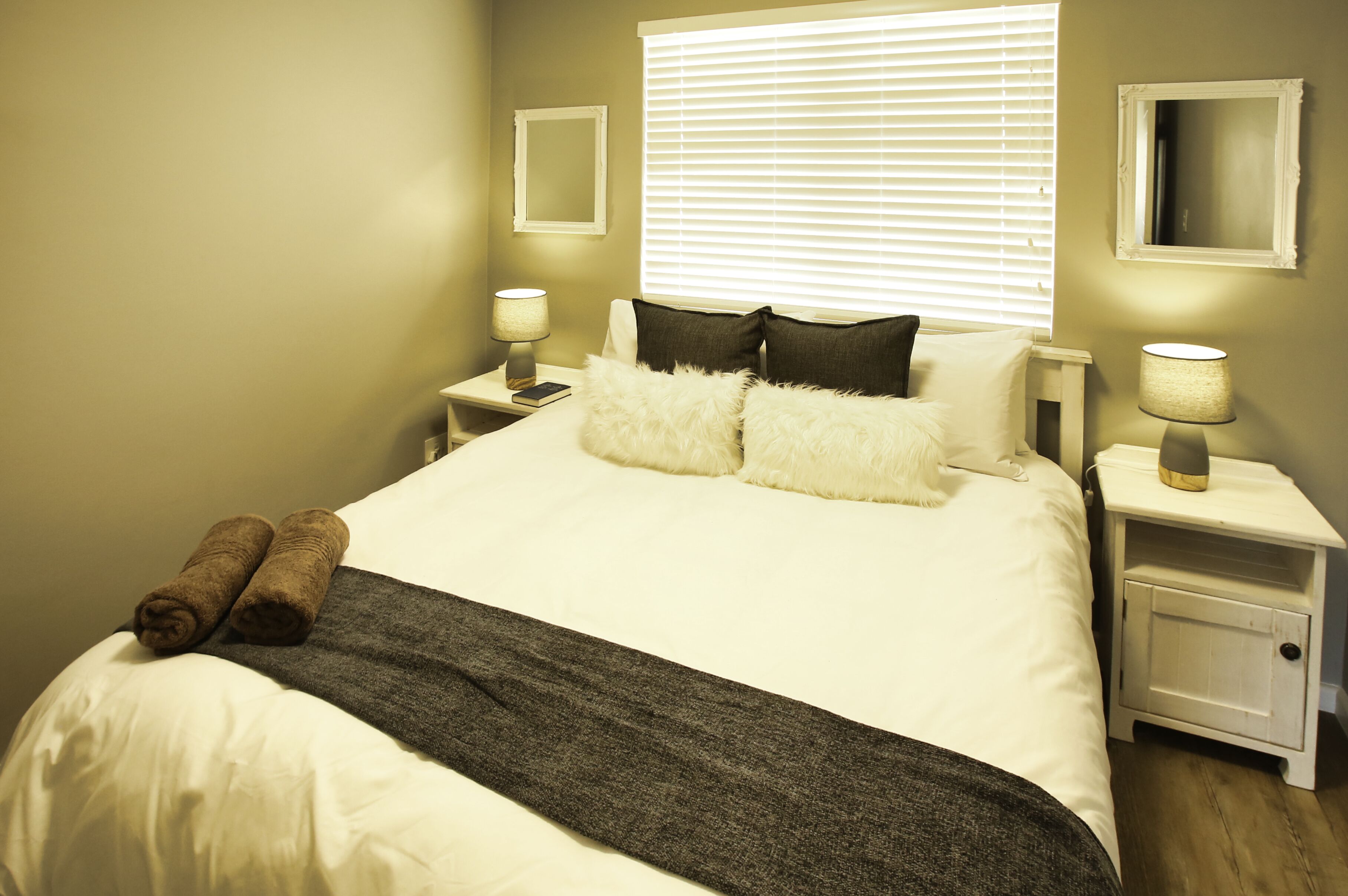 Una cama bien hecha con almohadas blancas y oscuras, una manta gris y dos toallas enrolladas a un lado. Hay dos mesitas de noche con lámparas y un espejo en la pared.