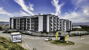 Un edifici d'apartaments modern amb múltiples balcons, una zona enjardinada amb una bandera i un carrer amb vehicles.