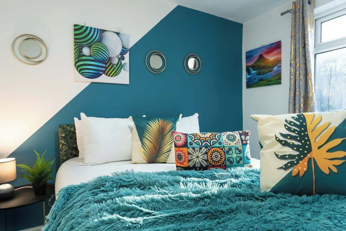 A bedroom with a bed, pillows, a blue wall, a painting, and a window with curtains.