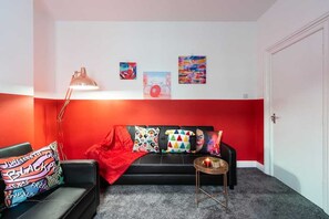 Una sala de estar moderna con un sofá negro, cojines coloridos, una manta roja, una lámpara de pie y decoración en la pared.
