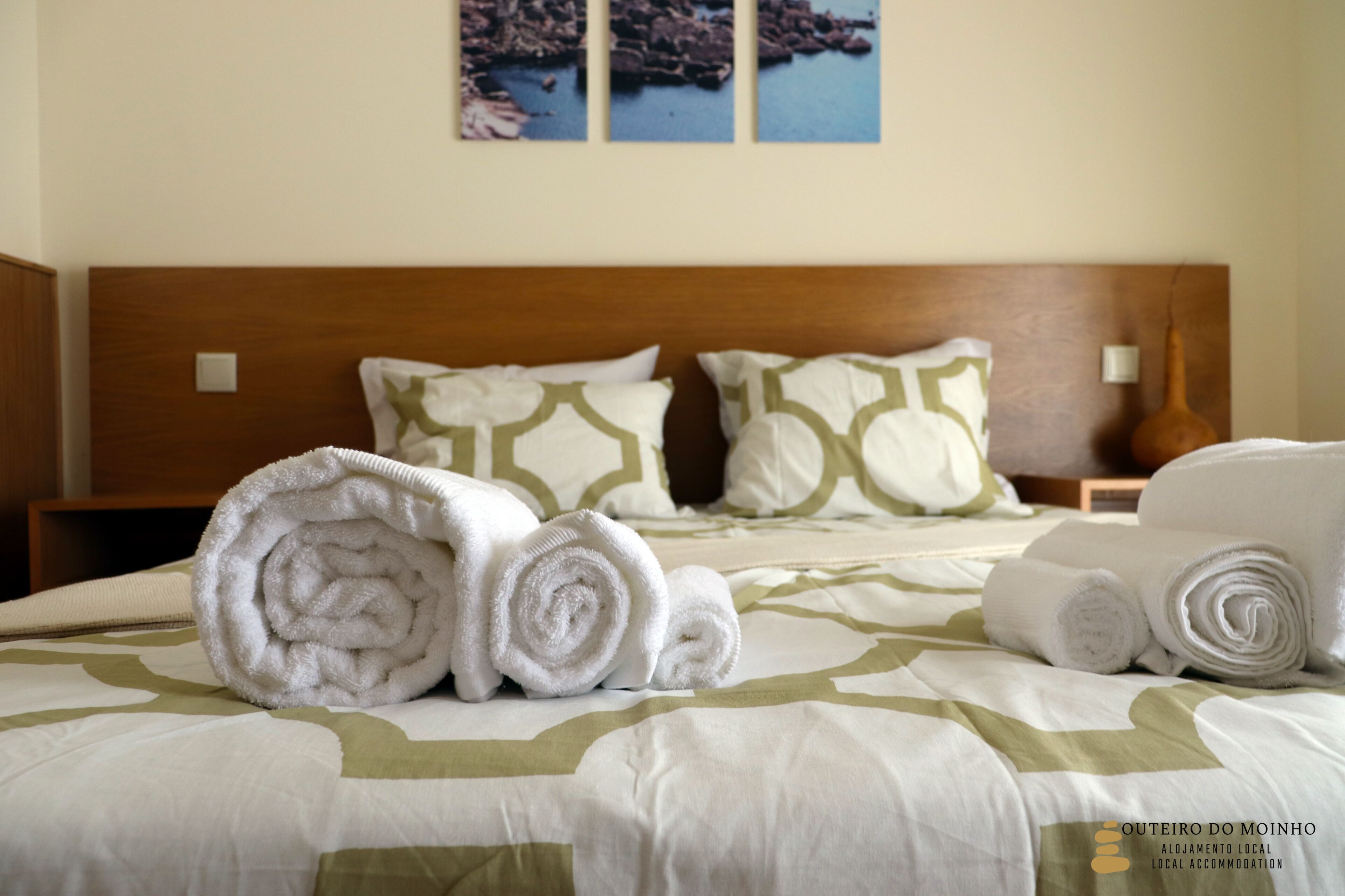 Una cama bien hecha con toallas enrolladas y almohadas estampadas, un cabecero de madera y una pared con cuadros.
